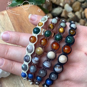 May include: Four macrame bracelets featuring round, multi-colored gemstone beads in various shades of blue, orange, green, brown, and white. The bracelets are crafted with white, brown, and black cord, and include a white hand-drawn planet graphic.