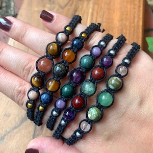 May include: Five macrame bracelets made with black cord, each featuring a series of round, multi-colored natural stone beads and small metallic spacers. The stones display various colors including brown, orange, green, purple, and clear.