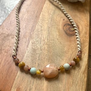 May include: A handmade necklace featuring a woven cord with a variety of beads. The necklace has a large, peach-colored stone in the center, surrounded by smaller beads in shades of yellow, pink, and blue. The necklace is displayed on a wooden surface.