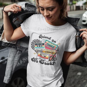 Puede incluir: Camiseta blanca con un gr&aacute;fico de cinta de cassette retro. El dise&ntilde;o presenta cintas de cassette coloridas con el texto "Raised On No Doubt". La camiseta la lleva una persona.