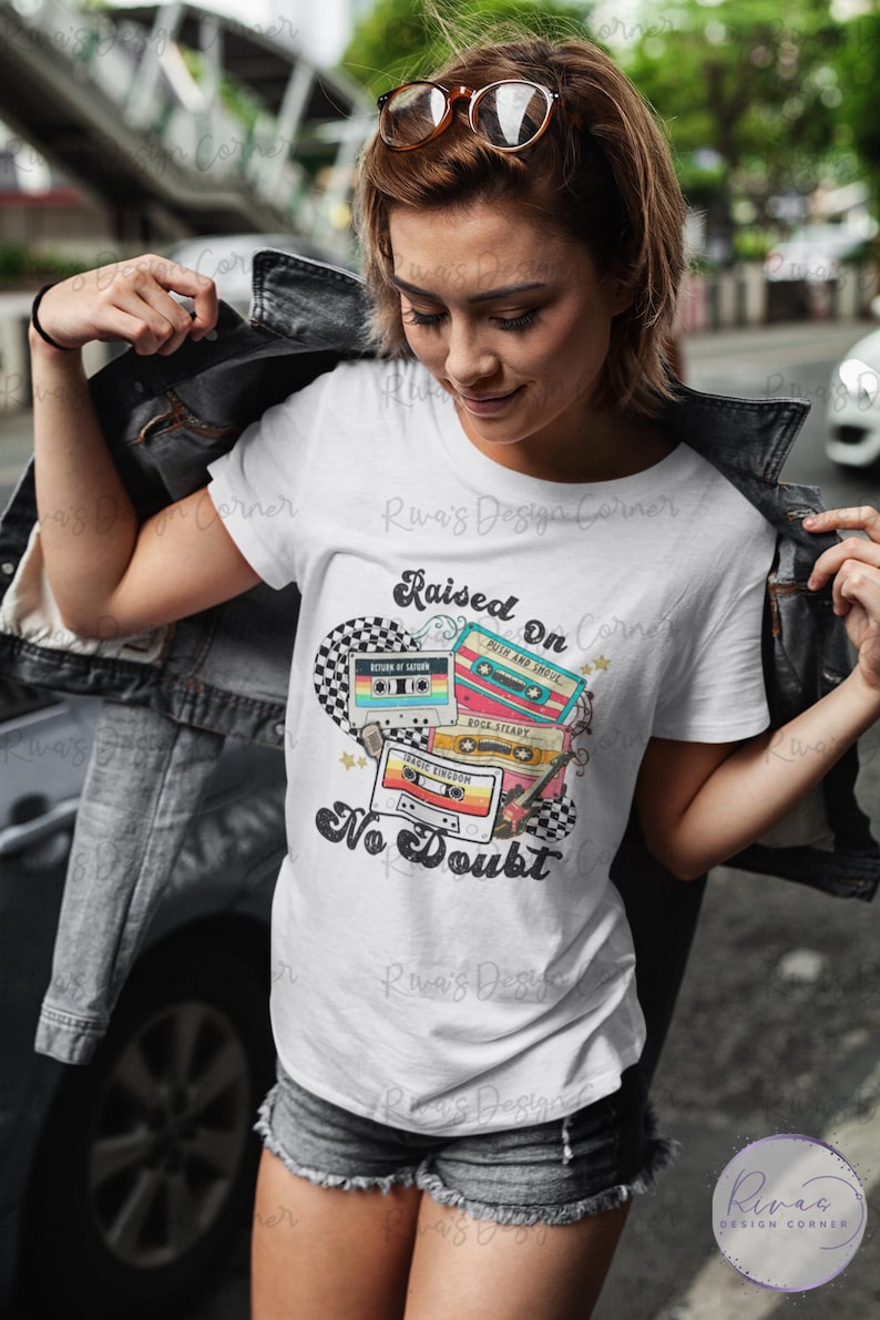 Puede incluir: Camiseta blanca con un gr&aacute;fico de cinta de cassette retro. El dise&ntilde;o presenta cintas de cassette coloridas con el texto "Raised On No Doubt". La camiseta la lleva una persona.