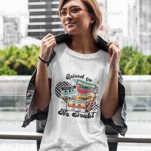 Puede incluir: Camiseta blanca con un dise&ntilde;o retro de casete y el texto "Raised On No Doubt". El dise&ntilde;o presenta cintas de casete coloridas y un fondo a cuadros. La persona lleva gafas y una chaqueta negra.