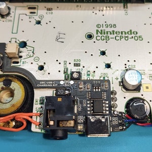 以下が含まれることがあります： さまざまな電子部品が取り付けられた回路基板のクローズアップ。基板には「&copy;1998 Nintendo CGB-CPU-05」と「GBC USB-C CHARGING KIT」の文字があります。スピーカー、ヘッドホンジャック、その他の小さな部品が見えます。