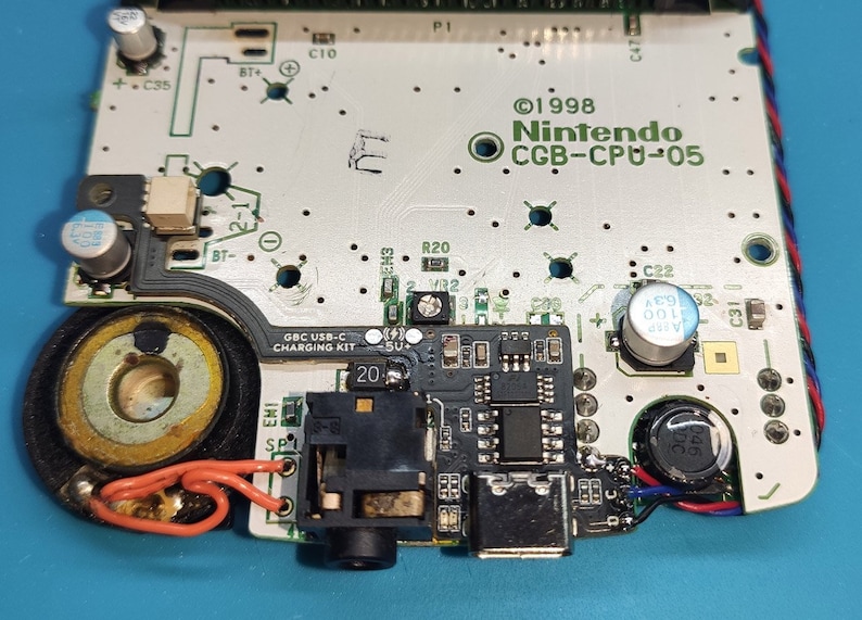 以下が含まれることがあります： さまざまな電子部品が取り付けられた回路基板のクローズアップ。基板には「&copy;1998 Nintendo CGB-CPU-05」と「GBC USB-C CHARGING KIT」の文字があります。スピーカー、ヘッドホンジャック、その他の小さな部品が見えます。