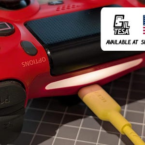 Könnte beinhalten: Ein roter Gamecontroller mit schwarzen Knöpfen und einem gelben Ladekabel. Auf dem Controller steht das Wort "OPTIONS". Ein weißes Rechteck mit dem Text "GIL TESA" und der amerikanischen Flagge befindet sich in der oberen rechten Ecke.