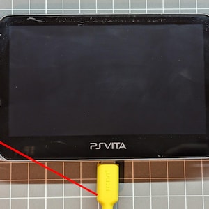 May include: Black Sony PlayStation Vita handheld game console with a bright screen, control buttons, and a yellow charging cable plugged in. The console has the text "PS VITA" printed on the front.