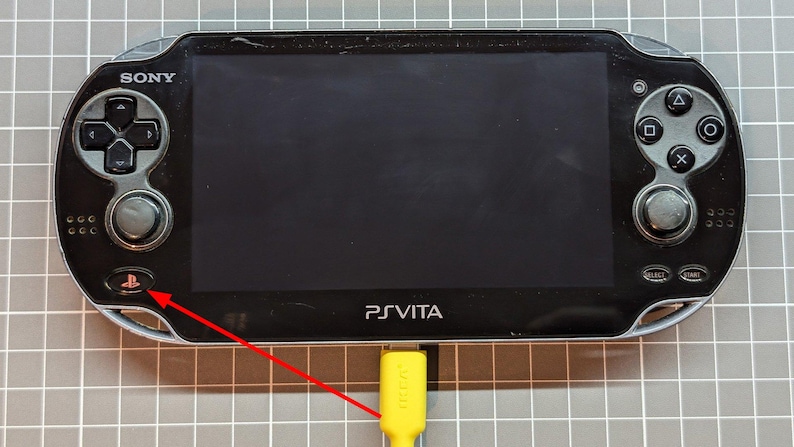 May include: Black Sony PlayStation Vita handheld game console with a bright screen, control buttons, and a yellow charging cable plugged in. The console has the text "PS VITA" printed on the front.