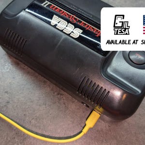 May include: Black Sega Master System II console with a yellow cable plugged in. The console has the text "Master System II" and "SEGA" on it. A white rectangle with the text "GIL TESA" and the American flag is in the upper right corner.