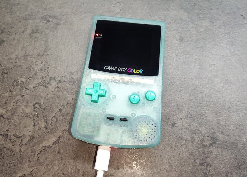 以下が含まれることがあります： 黒い画面とレインボーカラーで「GAME BOY COLOR」の文字が入った、ミントグリーンのゲームボーイカラー携帯ゲーム機。白い充電ケーブルでコンソールが接続されています。電源インジケーターが点灯しています。