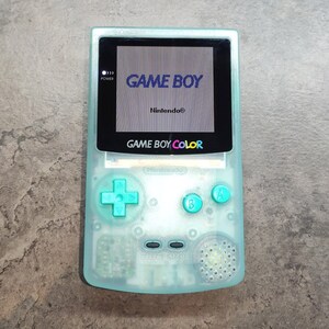 以下が含まれることがあります： 透明なティール色のゲームボーイカラー携帯ゲーム機。画面にはゲームボーイのロゴが表示されています。コンソールにはティール色のボタンと方向パッドがあります。「GAME BOY COLOR」の文字がコンソールに印刷されています。