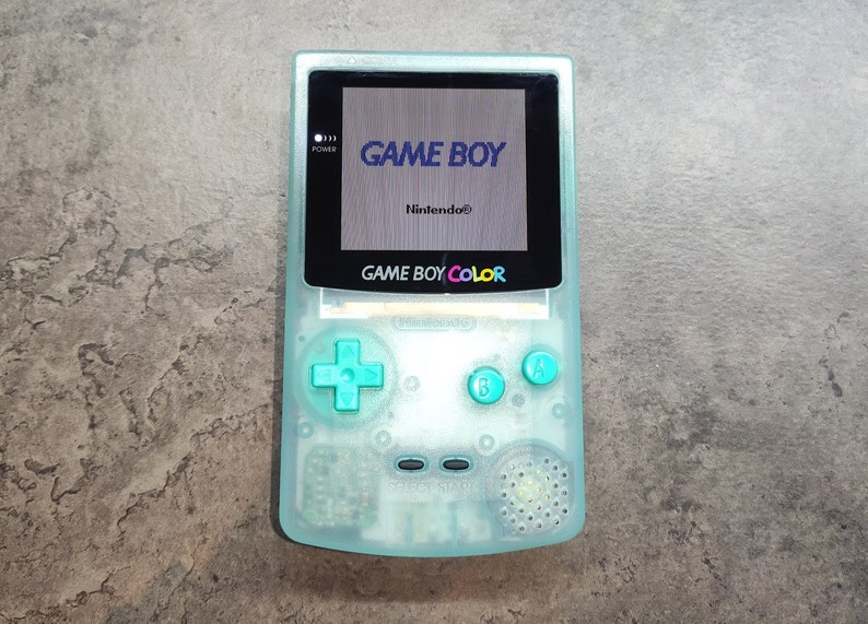 以下が含まれることがあります： 透明なティール色のゲームボーイカラー携帯ゲーム機。画面にはゲームボーイのロゴが表示されています。コンソールにはティール色のボタンと方向パッドがあります。「GAME BOY COLOR」の文字がコンソールに印刷されています。