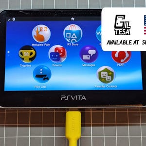 May include: Black Sony PS Vita handheld game console with a bright screen displaying various app icons. The console has a yellow charging cable plugged in. The screen shows the text "PS Vita".