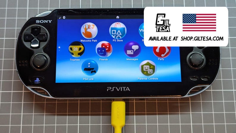 May include: Black Sony PS Vita handheld game console with a bright screen displaying various app icons. The console has a yellow charging cable plugged in. The screen shows the text "PS Vita".