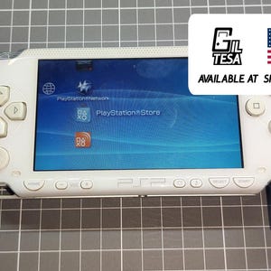 Puede incluir: Consola de juegos portátil blanca con una pantalla azul que muestra un menú. La consola tiene un panel direccional, botones y un cable de carga azul. La imagen incluye el texto "PlayStation Store".