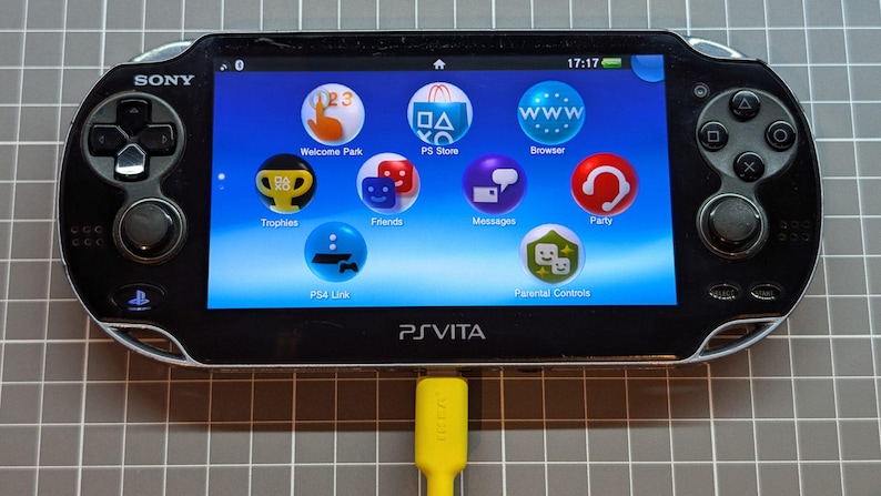 May include: Black Sony PS Vita handheld game console with a bright blue screen displaying app icons. The console has a yellow charging cable plugged in. The screen shows icons for Welcome Park, PS Store, Browser, Trophies, Friends, Messages, Party, PS4 Link, and Parental Controls.