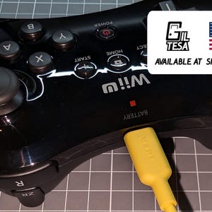 Könnte beinhalten: Schwarzes Wii U-Gamepad mit Tasten, Analog-Stick und gelbem Ladekabel. Das Gamepad hat das Wii U-Logo und den Text "AVAILABLE AT SHOP.GILTESA.COM" mit der amerikanischen Flagge.