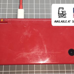 Puede incluir: Una consola de juegos portátil roja con un botón circular negro y un cable de carga enchufado. El dispositivo tiene forma rectangular y un acabado brillante. Una pegatina con el texto "GIL TESA" y la bandera estadounidense es visible.