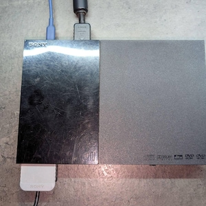 Puede incluir: Un reproductor de DVD Sony plateado y gris con un adaptador de corriente y cables enchufados. El reproductor de DVD tiene el logotipo de Sony y símbolos de DVD en el lado derecho.