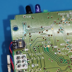 以下が含まれることがあります： さまざまな電子部品を備えた緑色の回路基板のクローズアップ。基板には、トレース、はんだ付けポイント、テキストラベルがあります。2つの小さなカラーLEDライトが見えます。ワイヤー付きの小さな黒いコンポーネントが取り付けられています。