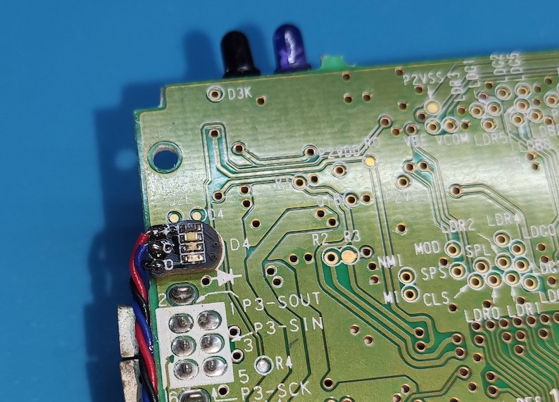 以下が含まれることがあります： さまざまな電子部品を備えた緑色の回路基板のクローズアップ。基板には、トレース、はんだ付けポイント、テキストラベルがあります。2つの小さなカラーLEDライトが見えます。ワイヤー付きの小さな黒いコンポーネントが取り付けられています。