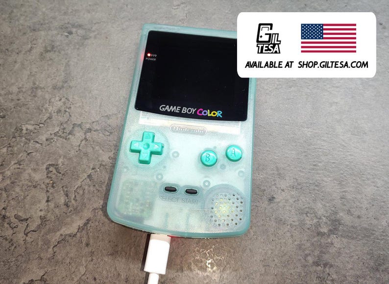 以下が含まれることがあります： ミントグリーンのゲームボーイカラー携帯ゲーム機。電源が入っており、充電中です。画面には黒い画面が表示されています。コンソールには十字型の方向パッドとカラーボタンがあります。「GAME BOY COLOR」の文字が表示されています。