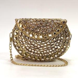 Könnte beinhalten: Eine halbkreisförmige goldfarbene Clutch mit einer komplexen Oberfläche, die mit klaren, facettierten und quadratischen Glassteinen besetzt ist. Die Tasche verfügt über einen goldfarbenen Metallkettenriemen und einen geschwungenen Klappenverschluss.