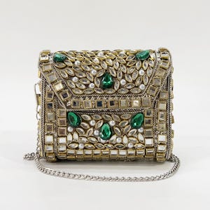 May include: A rectangular clutch bag featuring an intricate gold-tone metal frame adorned with numerous clear glass crystals, small white faux pearls, and several large, teardrop-shaped emerald green stones. It includes a silver-tone chain strap.