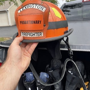 May include: An orange firefighter helmet with a black strap and a leather patch that reads "LEXINGTON PROBATIONARY FIREFIGHTER". The helmet is hanging from a black strap in the back of a truck.
