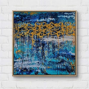 Può includere: Dipinto astratto in cornice di legno. L'opera presenta una superficie strutturata con sfumature di blu, turchese e bianco. Accenti dorati e neri creano un effetto a strati, con gocciolamenti e motivi lineari. Il dipinto quadrato è montato su un muro di mattoni bianchi.