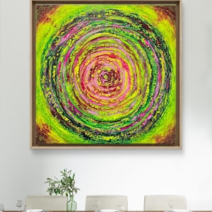 Può includere: Dipinto astratto con un motivo a spirale nei toni del verde neon, rosa e giallo, incorniciato in una cornice di legno marrone chiaro. L'opera è esposta sopra un tavolo da pranzo con sedie bianche e un vaso di piante verdi.