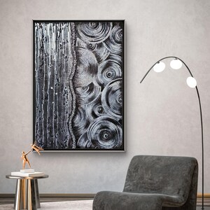 May include: Abstract black and white artwork in a black frame. The painting features a textured design with vertical drips on the left and concentric circles on the right. A modern gray chair and a gold side table with a sculpture are in the foreground.