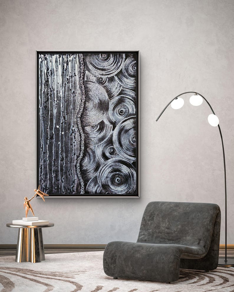 May include: Abstract black and white artwork in a black frame. The painting features a textured design with vertical drips on the left and concentric circles on the right. A modern gray chair and a gold side table with a sculpture are in the foreground.
