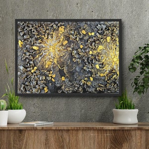 Può includere: Opera d'arte astratta in cornice nera. L'opera presenta una superficie strutturata con elementi neri, grigi e dorati. Gli accenti dorati si irradiano verso l'esterno, creando un contrasto sorprendente. L'opera è esposta su una superficie di legno.