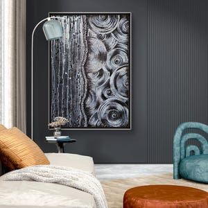 May include: Abstract black and white artwork with a textured, layered design. The painting is framed and hangs on a dark gray wall. A white floor lamp with a blue shade illuminates the artwork. A beige sofa with orange pillows and a brown ottoman are in the foreground.