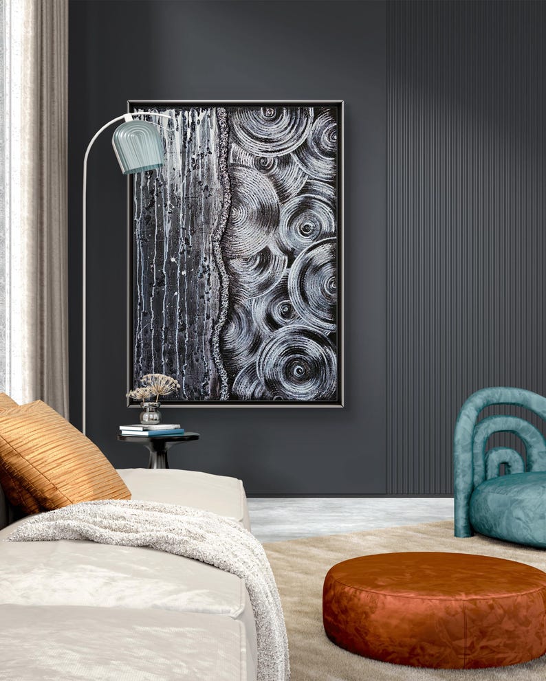 May include: Abstract black and white artwork with a textured, layered design. The painting is framed and hangs on a dark gray wall. A white floor lamp with a blue shade illuminates the artwork. A beige sofa with orange pillows and a brown ottoman are in the foreground.