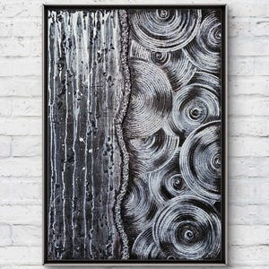 May include: Abstract black and white artwork in a black frame. The left side features vertical streaks of white paint over a dark background. The right side displays concentric circles in white and gray. The artwork is mounted on a white brick wall.