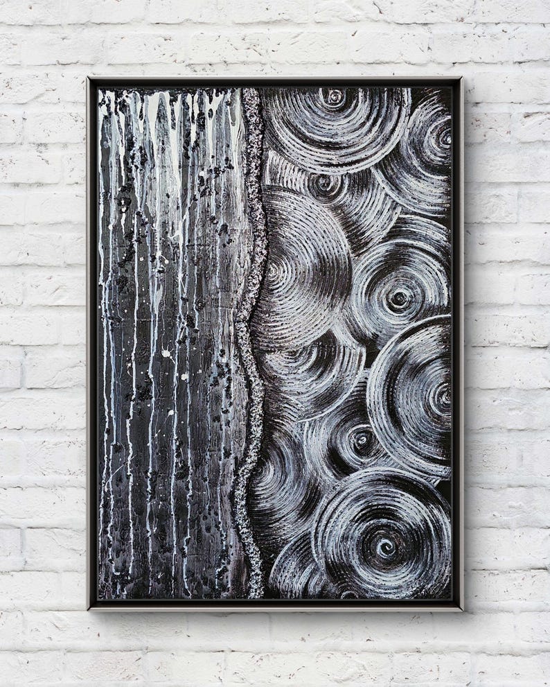 May include: Abstract black and white artwork in a black frame. The left side features vertical streaks of white paint over a dark background. The right side displays concentric circles in white and gray. The artwork is mounted on a white brick wall.