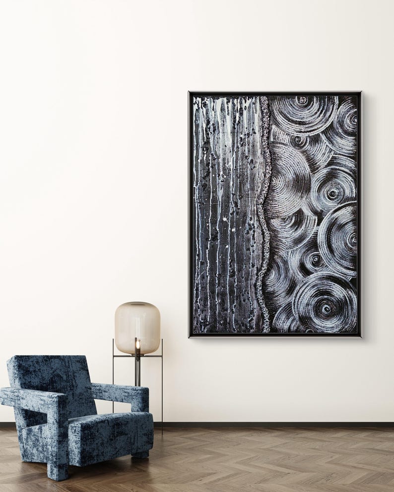 May include: Abstract black and white artwork in a black frame. The painting features a vertical section with white drips and a textured side with concentric circles. A blue velvet armchair and a lamp are in the foreground.