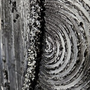 May include: Close-up of a textured abstract artwork in shades of gray and black. The piece features a swirling pattern on the right, a rough, granular texture in the center, and a vertical, wood-grain-like texture on the left. The artwork has a three-dimensional appearance.