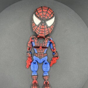 May include: A 3D-printed Spider-Man action figure. The figure features a red and black head with white eye details, a red and blue torso, and articulated limbs. The figure is posed upright on a gray surface.