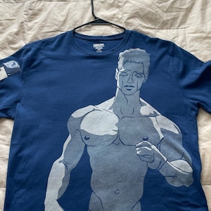 Può includere: Una maglietta blu con una stampa grafica bianca di un torso maschile. La grafica è un disegno a linee di un uomo muscoloso con un'espressione triste sul viso. La maglietta ha un'etichetta sulla manica che dice "SHIVAIZE 15 €".