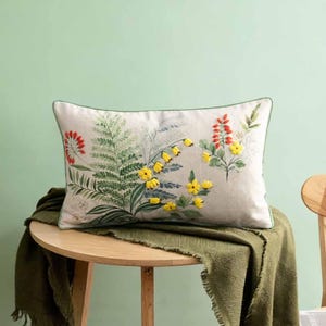 May include: Rectangular cushion with a floral design. The cushion features embroidered yellow, red, and green flowers and foliage on a cream background. It has a green border and is placed on a wooden table.