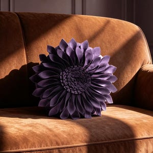 May include: A round, purple, flower-shaped decorative pillow with layered, petal-like fabric folds sits centered on a brown, velvet-textured sofa. The sofa features a smooth, upholstered surface with piped edges and dark, rounded wooden legs.