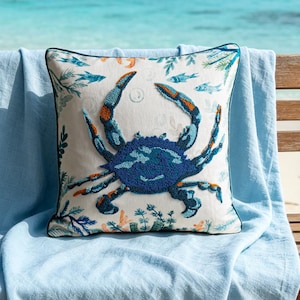 Op de afbeelding: Een vierkant decoratief kussen met een blauw krabontwerp, rustend op een lichtblauwe deken op een houten bank. De krab is voornamelijk blauw met oranje accenten. Het kussen heeft een witte achtergrond met vis- en koraalmotieven.