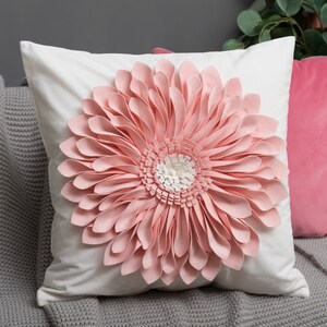 May include: A white pillow with a large pink flower design. The flower is made of fabric petals and has a white center. The pillow is sitting on a gray knitted throw blanket.