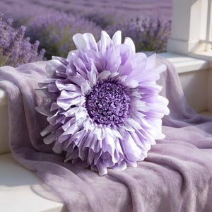 Könnte beinhalten: Ein rundes Dekokissen in Blumenform mit geschichteten hellvioletten und lavendelfarbenen Blütenblättern. Die Mitte des Kissens weist eine dichte, gerüschte Textur in einem dunkleren Violettton auf. Darunter liegt ein passender hellvioletter Stoff.