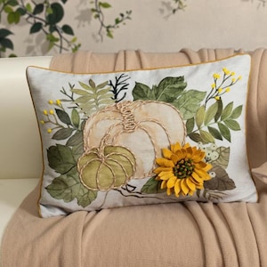 May include: Rectangular decorative pillow featuring a pumpkin and sunflower design. The pillow has a light background with a gold trim. The pumpkin is beige and green, and the sunflower is yellow. The pillow is on a beige blanket.