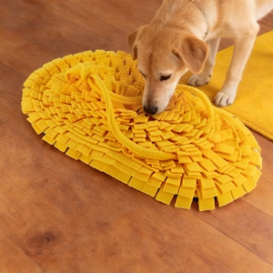 Könnte beinhalten: Eine gelbe Hunde-Puzzlematte mit strukturierter Oberfläche, die den Geruchssinn des Hundes anregen und die Futtersuche fördern soll. Ein hellbrauner Hund schnuppert an der Matte. Die Matte ist oval und aus weichem Material.