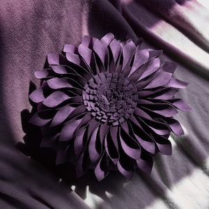 May include: A round, deep purple decorative pillow featuring layered, petal-like fabric strips arranged in a dense, circular flower pattern. The center consists of smaller, upright rectangular fabric pieces, while the outer edges feature longer, curved, folded petals.