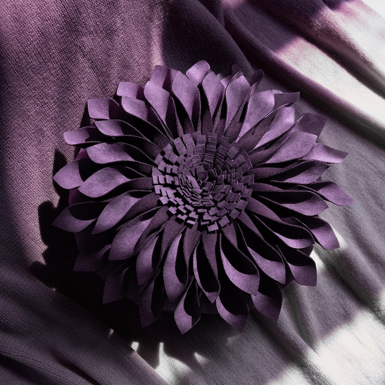 May include: A round, deep purple decorative pillow featuring layered, petal-like fabric strips arranged in a dense, circular flower pattern. The center consists of smaller, upright rectangular fabric pieces, while the outer edges feature longer, curved, folded petals.